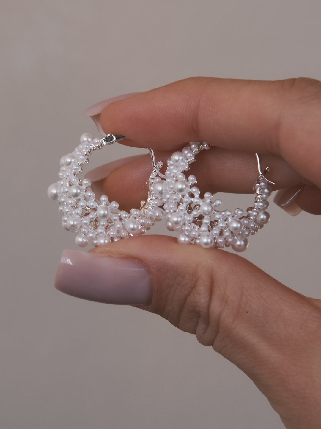 Elderflower Huggies in silver-tone, a pair of openwork huggies full of cream-white beads, held between two fingers against a pale grey background