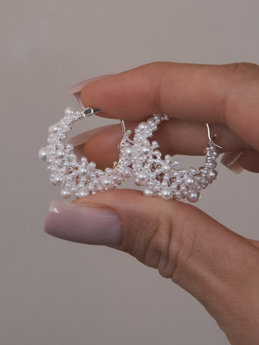 Elderflower Huggies in silver-tone, a pair of openwork huggies full of cream-white beads, held between two fingers against a pale grey background